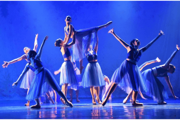 "Bailarinos no palco do Festival de Dança de Joinville em meio a uma apresentação vibrante, com trajes coloridos e coreografia sincronizada, sob iluminação cênica. Plateia lotada aparece ao fundo, demonstrando a grandiosidade do maior festival de dança do mundo."