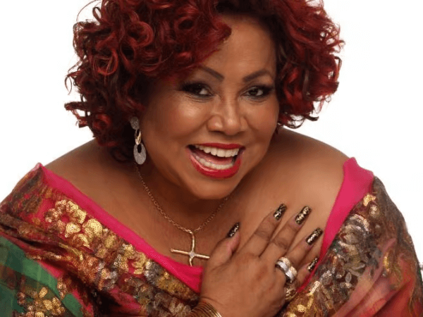 "Alcione, conhecida como 'A Voz do Samba', em performance no palco. Seu vestido colorido e expressão apaixonada refletem sua energia característica, enquanto canta com o microfone próximo ao rosto."