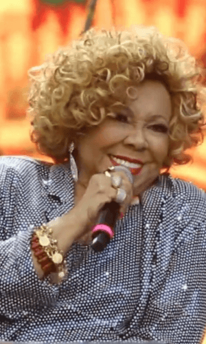 "Alcione Nazareth, a cantora e ícone do samba brasileiro, sorrindo durante apresentação musical. Veste traje elegante e segura microfone, com palco iluminado ao fundo."